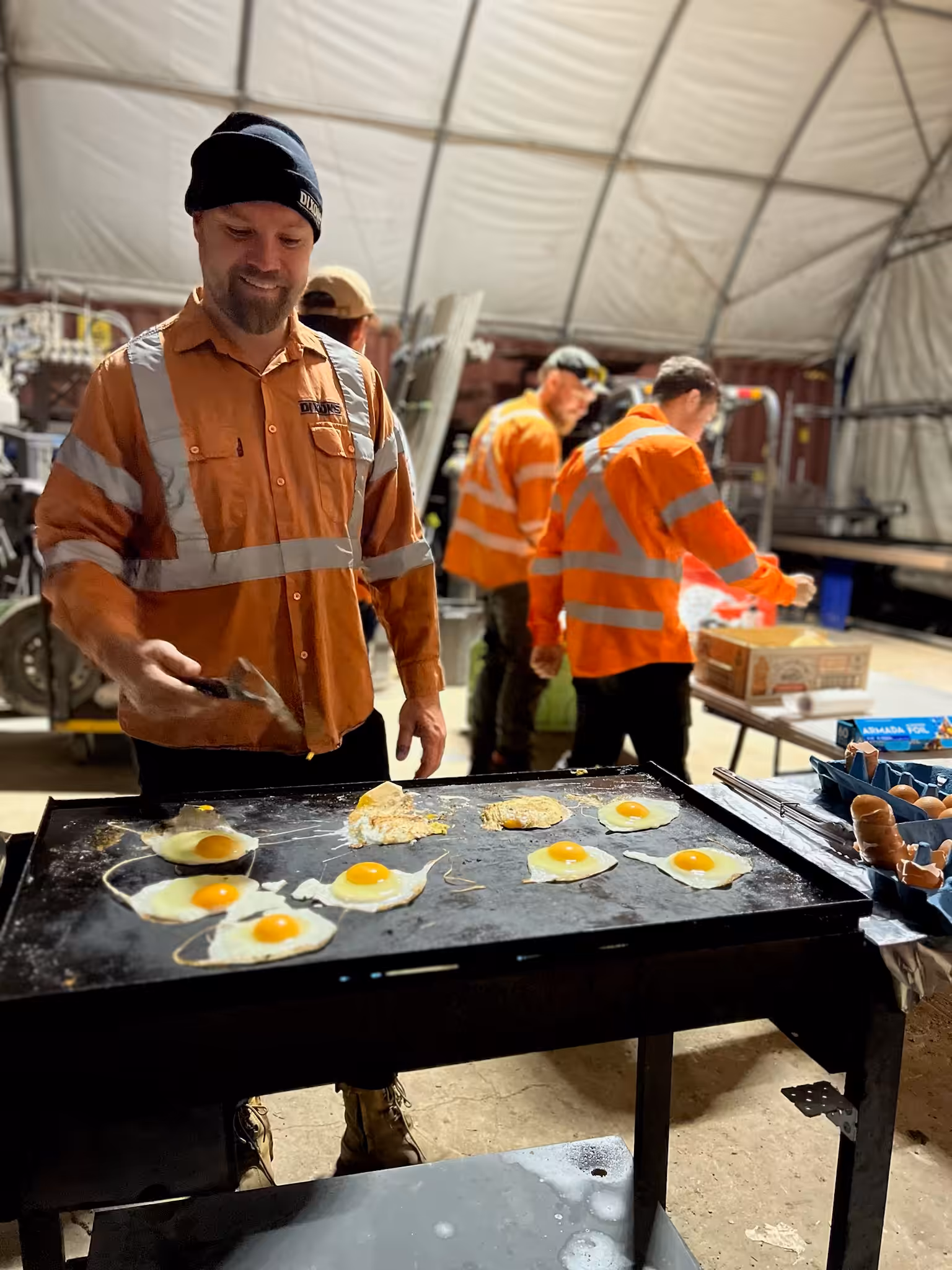 Construction workers cooking eggs on large griddle indoors